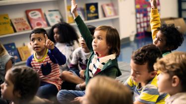 Share your ideas! Grade 1 student with hand raised among group of classmates seated on floor.