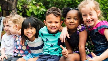 Kindergarten Registration Six smiling, diverse pre-school children seated outdoors with arms draped around one another's shoulders.