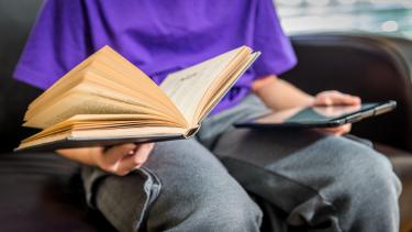 Child in purple shirt and grey jeans sitting holding an open book in one hand and tablet in the other.